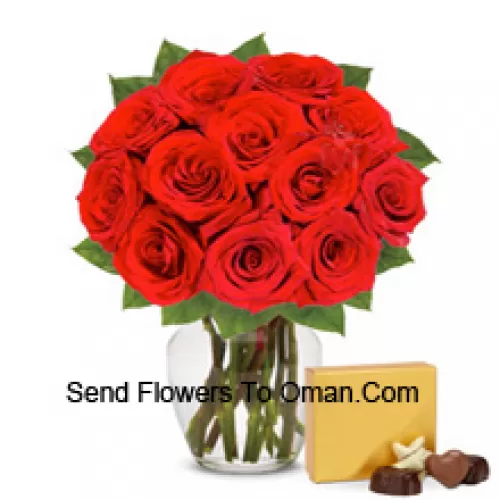 12 punaista ruusua muutamien saniaisten kera lasimaljakossa, mukana tuontisuklaarasia
