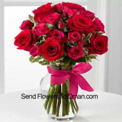 18 punaista ruusua kausittaisilla täytteillä lasimaljakossa, joka on koristeltu vaaleanpunaisella nauhalla